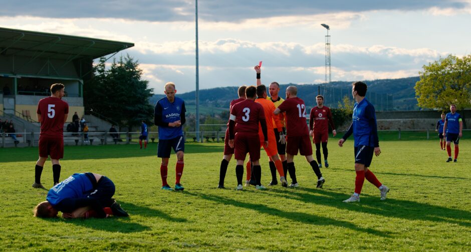 Sombat Penalty Sequence 14: Sombat lies on the ground in pain as referee Markus Kouba faces down Julius Pentek, Markus Lackner and Peter Gallus. Curiously in Premiere League football the rules were changed in 2016 to allow only a yellow card if a penalty kick is awarded on a DOGSA foul. Julius Pentek's mistake here was to shout at the referee. If he's kept quiet it might only have been a yellow card and a penalty kick. Subject: soccer;football;burgenland;kittsee;Jozef Sombat;Julius Pentek;Andreas Roth