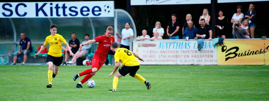 Sombat splits defenders again: Jozef Sombat slips past Mario Katzler again while substitute midfielder Florian Gombay chases the play. Kittsee coach Manfred Wachter observes the action from the bench, while Kittsee home bench fans enjoy a lead for a change this season. Subject: soccer;football;burgenland;kittsee;ASV Jahrndorf;Florian Gombay;Jozef Sombat;Mario Katzler;Manfred Wachter